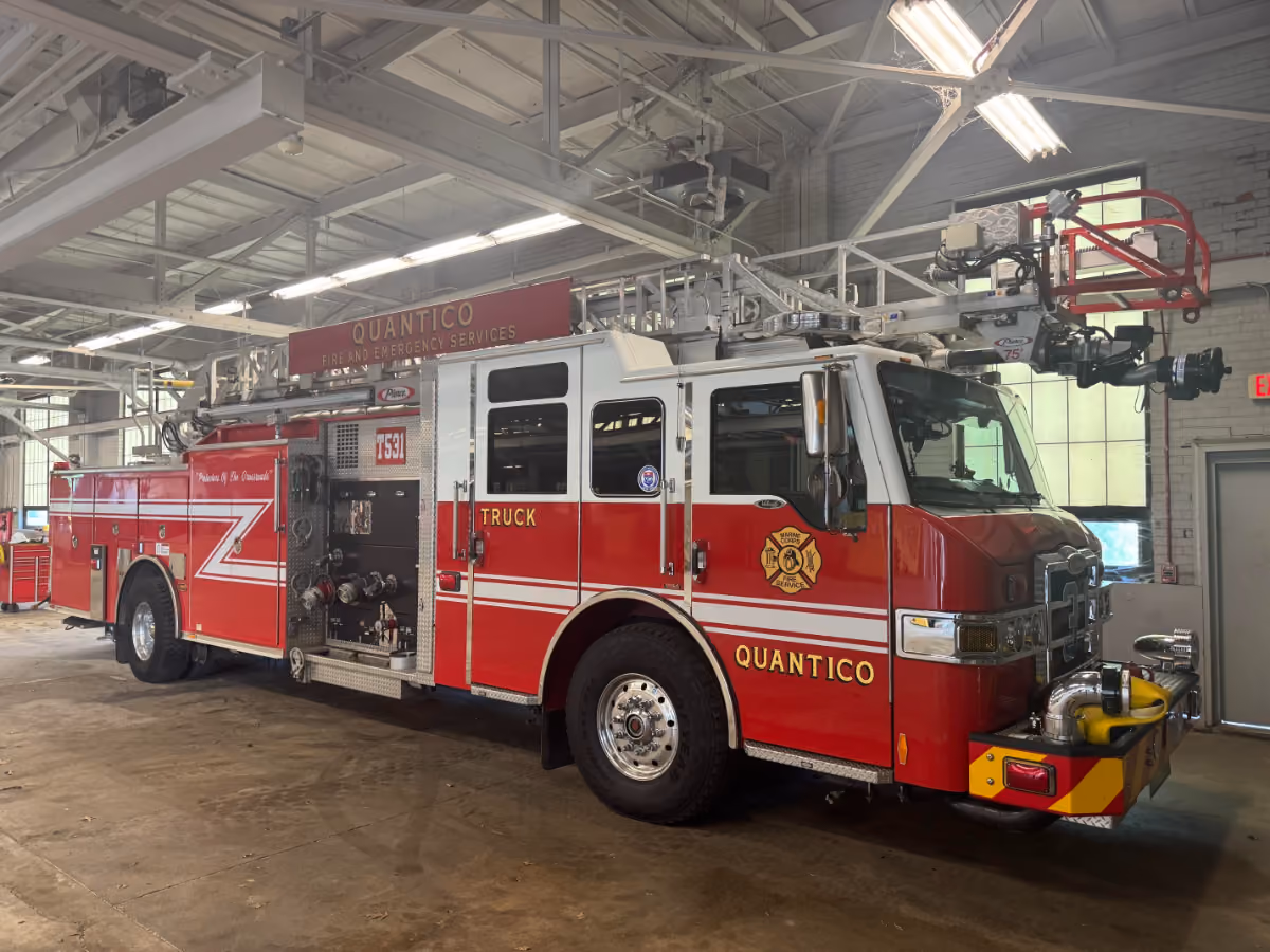 A red and white fire truck labeled "QUANTICO FIRE AND EMERGENCY SERVICES" is parked inside a fire station with high ceilings and fluorescent lights. The vehicle, identified as "Truck 75," features an extended ladder, multiple hoses, and department insignia.