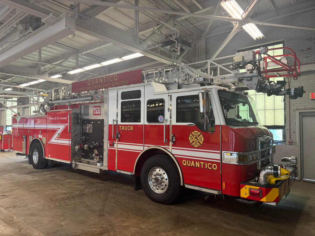 A red and white fire truck labeled "QUANTICO FIRE AND EMERGENCY SERVICES" is parked inside a fire station with high ceilings and fluorescent lights. The vehicle, identified as "Truck 75," features an extended ladder, multiple hoses, and department insignia.