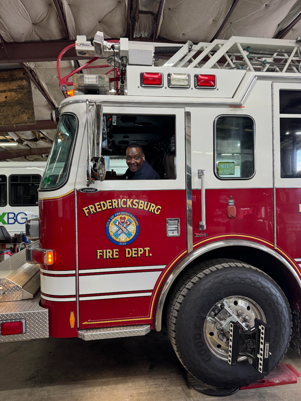 A smiling man is seated inside the driver’s seat of a red Fredericksburg Fire Department fire truck parked inside a garage with an exposed, insulated ceiling. The truck features a visible emblem, emergency lights, and a large front tire with a wheel lock.