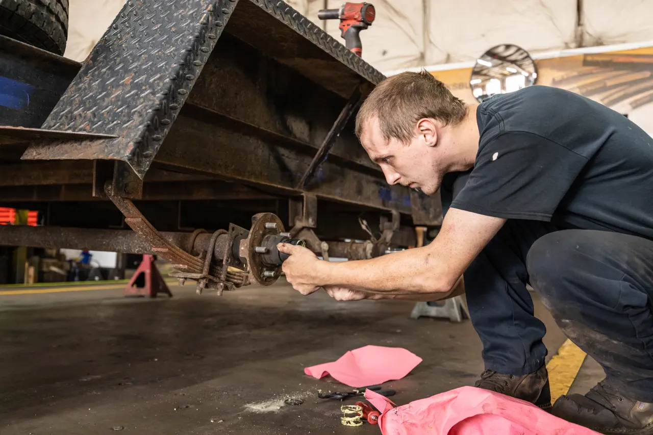 Trailer repair technician servicing a trailer axle and hub assembly with the wheel removed inside a heavy-duty repair shop.