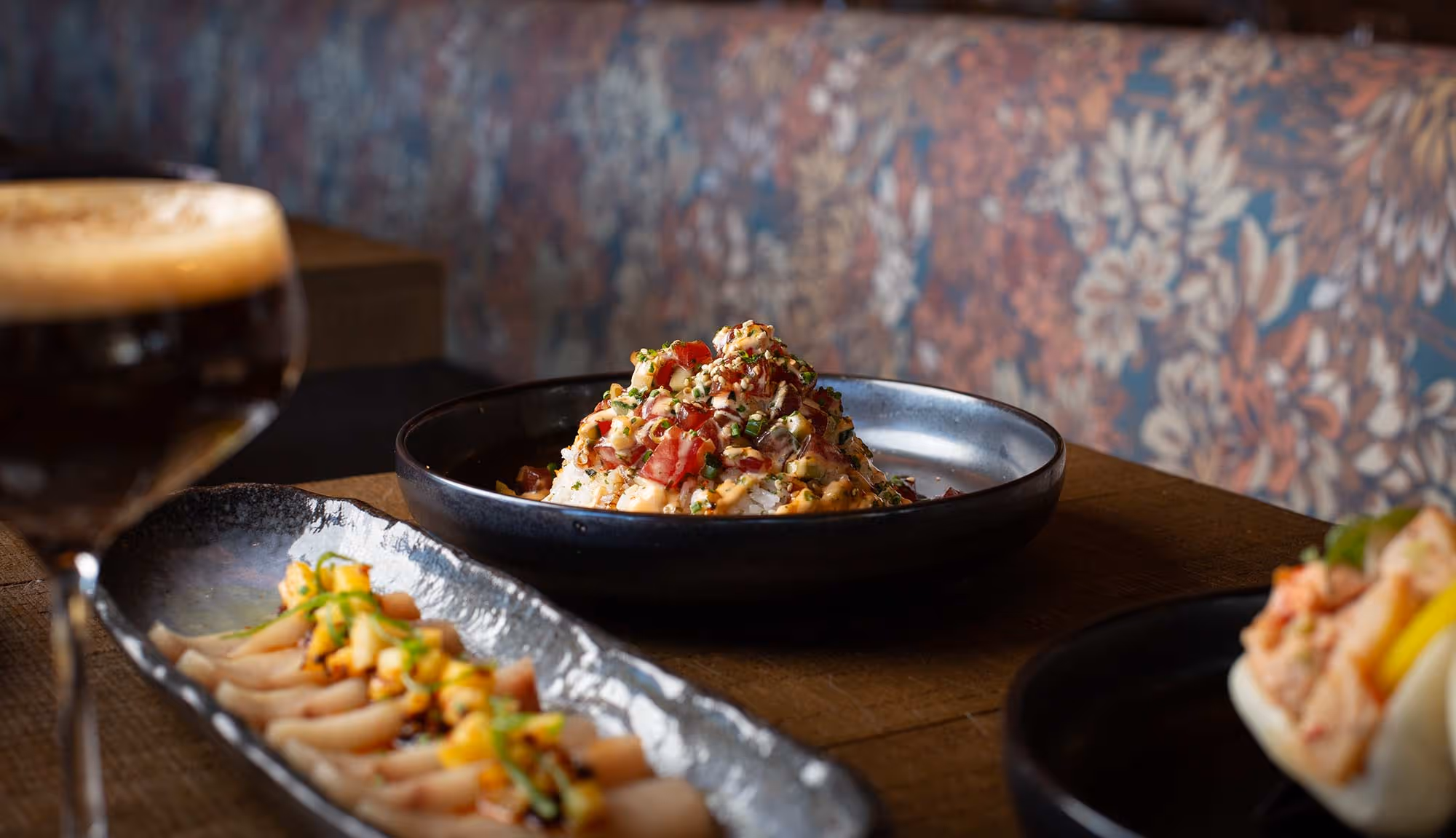 Close-up of a gourmet dining table featuring a poke bowl topped with diced tuna and garnishes, a plate of thinly sliced sashimi with mango salsa, a steamed bao bun, and a dark espresso cocktail, all set against a patterned upholstered booth in a stylish restaurant.