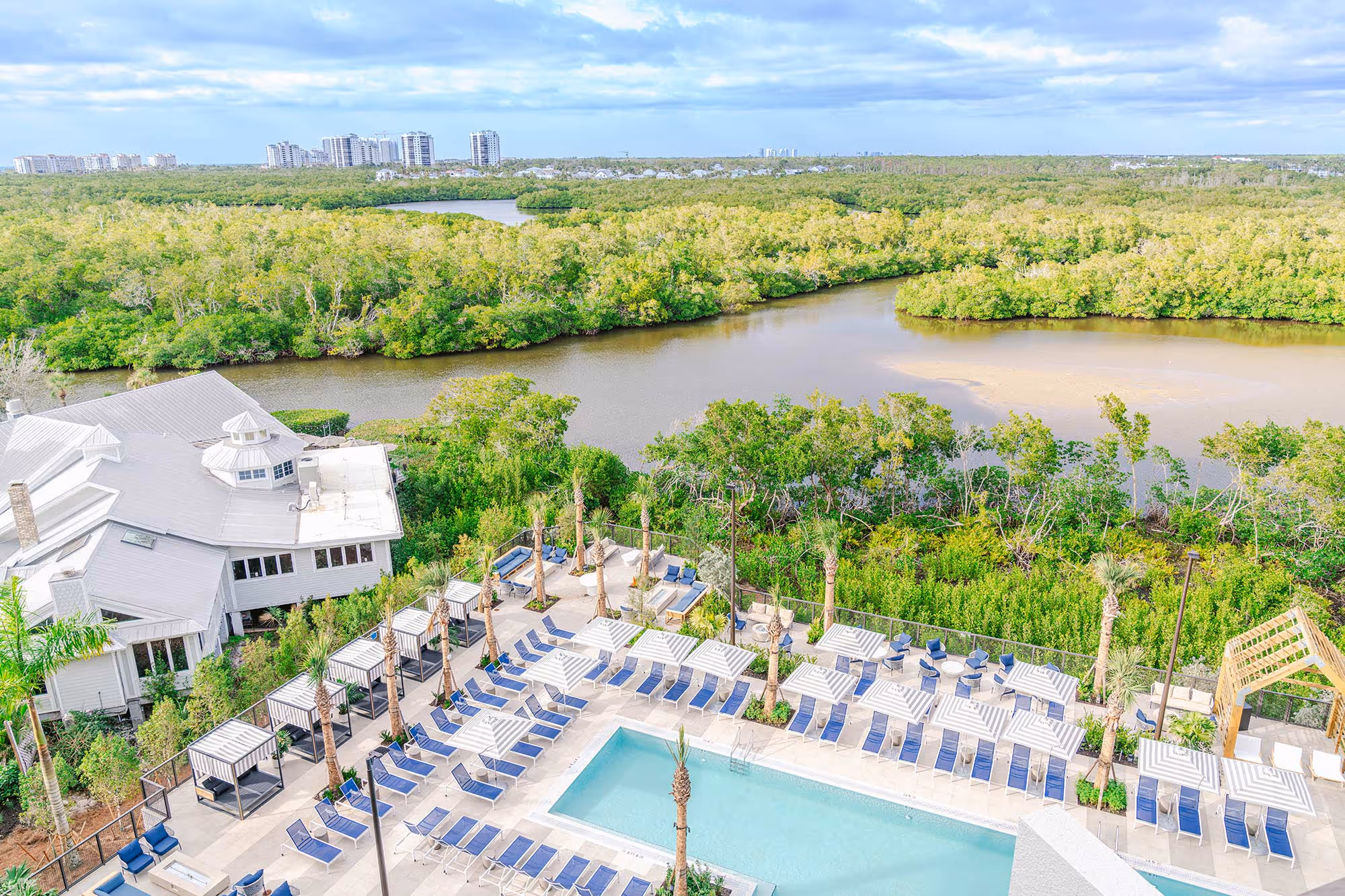 Overhead view of resort-style pool deck with striped cabanas, palm trees, and a winding river below.