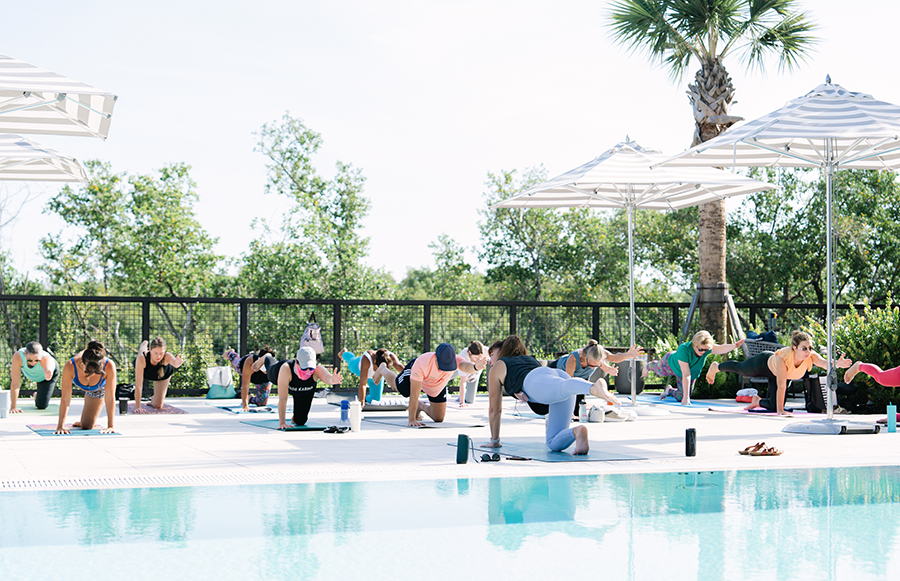 Group of people doing a yoga or fitness class on mats by a pool under white umbrellas.
