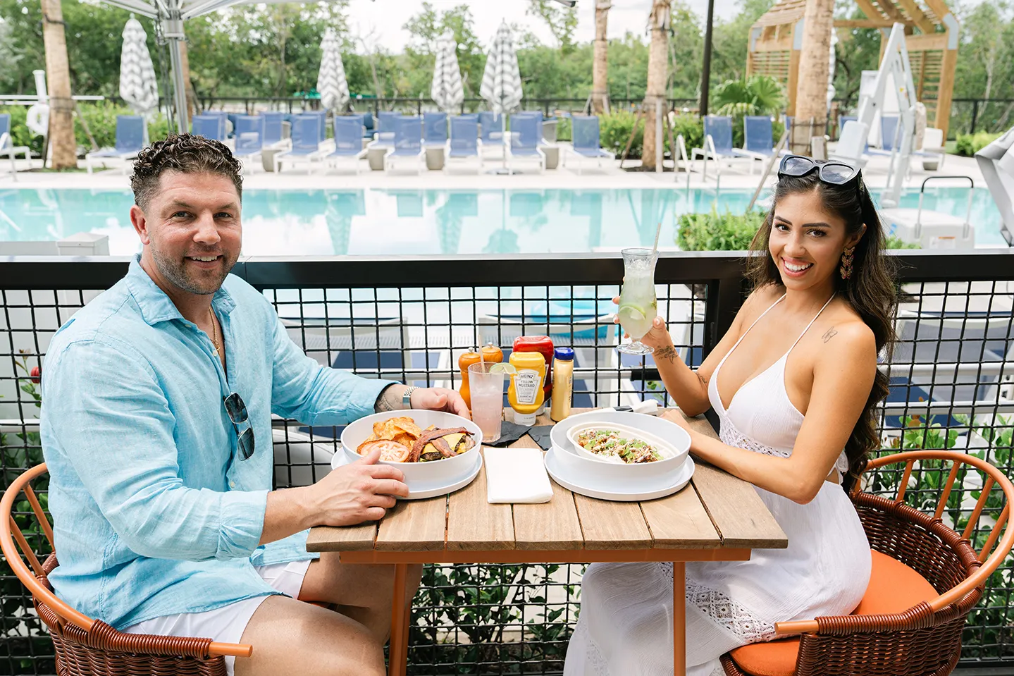 Man and woman smiling at a poolside table with plates of food and drinks, surrounded by greenery and lounge chairs.