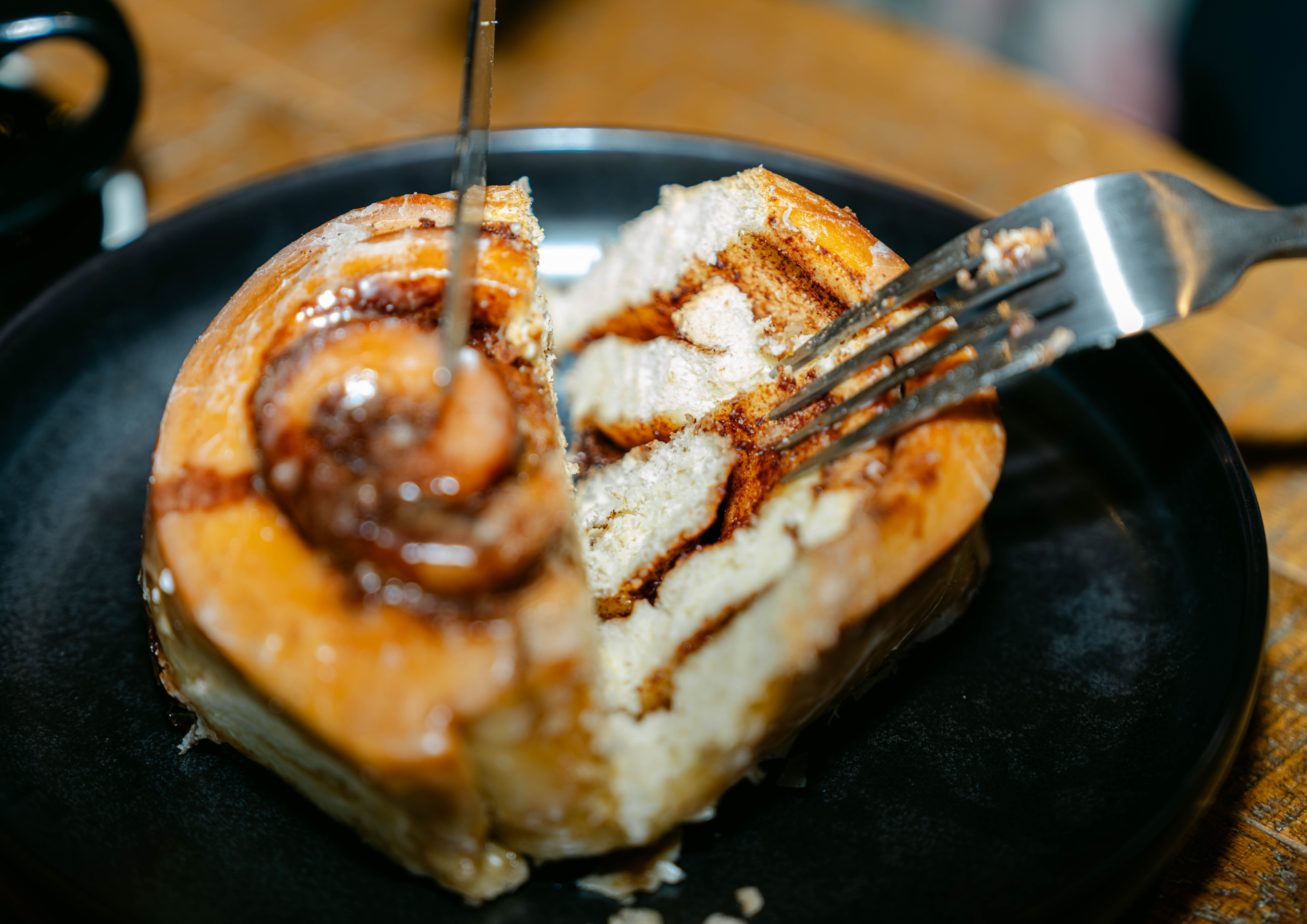 Fork cutting into a cinnamon roll on a black plate on a wooden table.