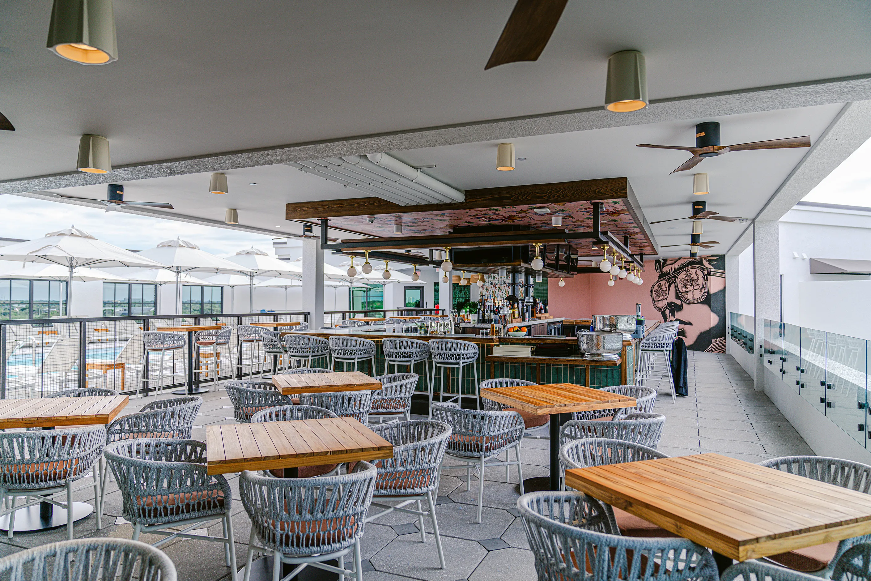 Modern outdoor rooftop bar with wooden tables, gray woven chairs, ceiling fans, and white umbrellas by a pool.