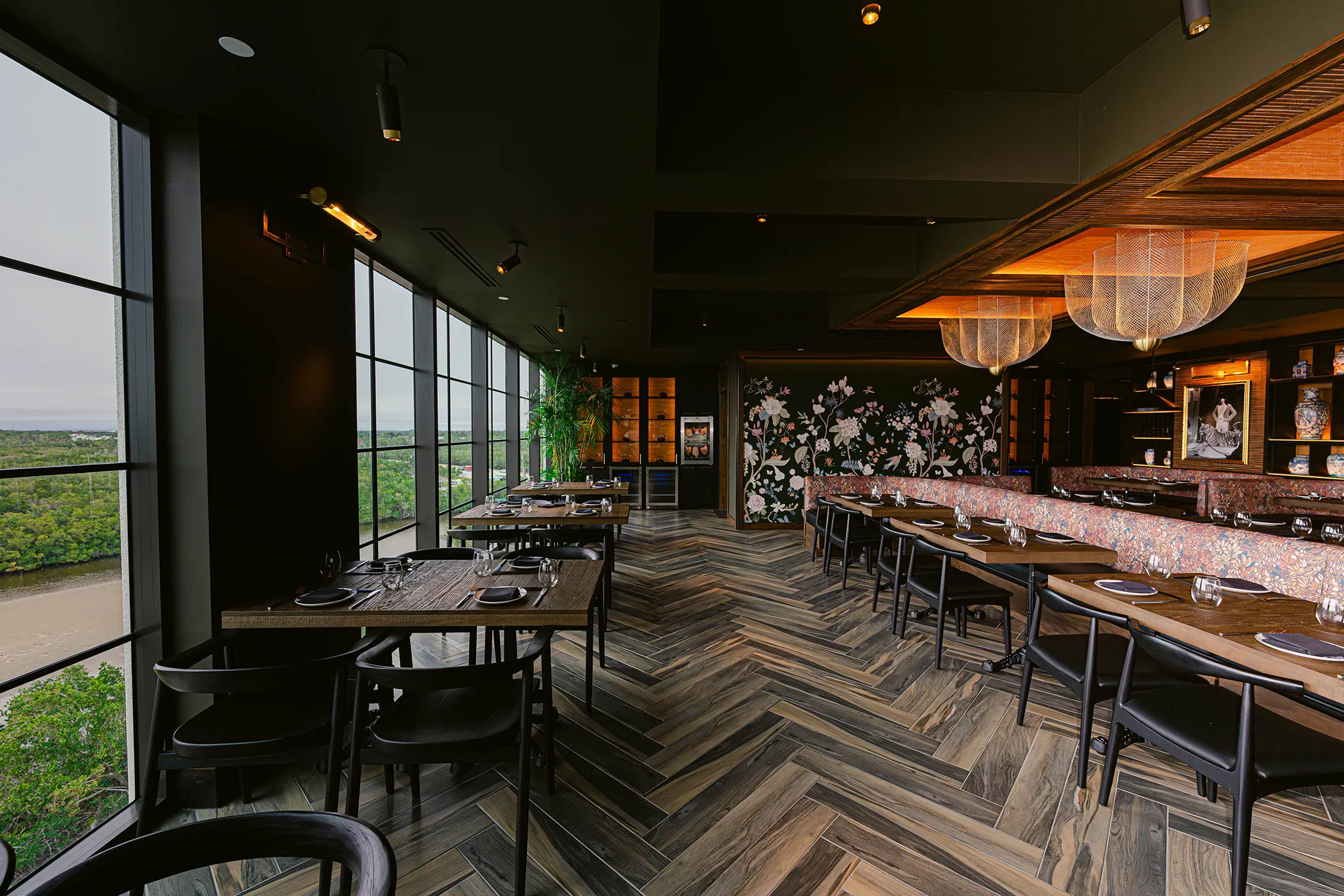 Upscale restaurant interior with wooden chevron floor, large windows overlooking greenery, floral wallpaper, and neatly set tables.
