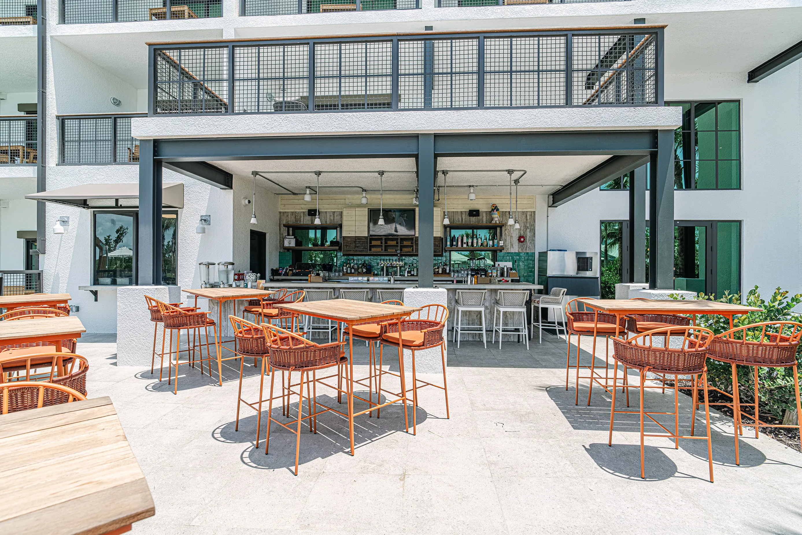 Outdoor bar area with orange wicker high chairs and wooden tables under a covered patio in a modern white building.