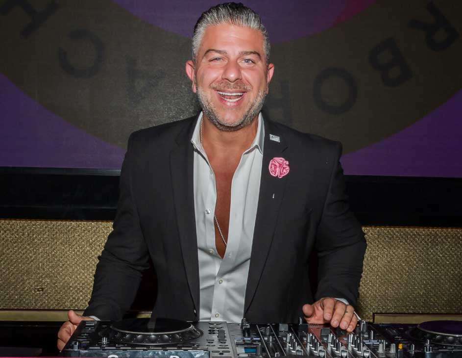Smiling man with slicked-back gray hair wearing a black suit and open white shirt standing behind DJ equipment.