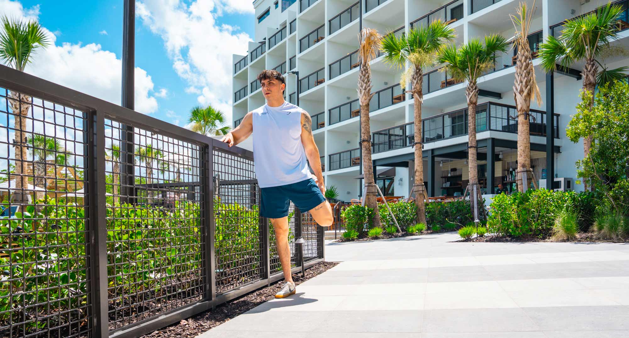 Man in athletic wear stretching his leg outdoors near a modern building with palm trees.