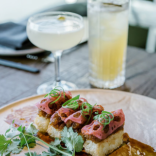 Plate with three pieces of sushi topped with slices of seared beef and garnished with green onions and cilantro, next to a cocktail in a coupe glass and a tall glass of a pale yellow drink with ice.