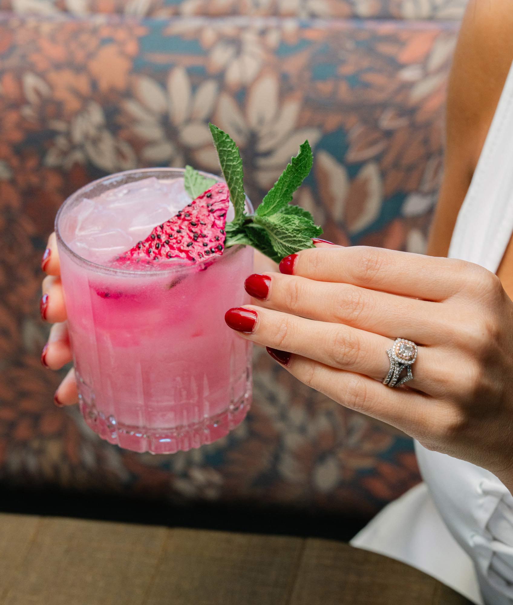 Hand with a diamond ring and red nail polish holding a pink iced drink garnished with a slice of dragon fruit and a sprig of fresh mint in a clear glass.