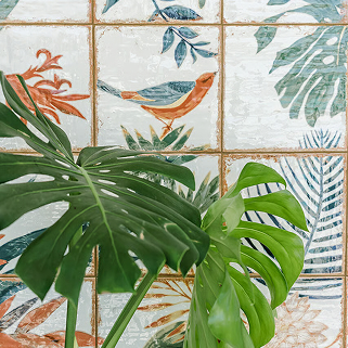 Green monstera leaves in front of a tiled wall depicting painted birds, blue and green leaves, and abstract plant designs.