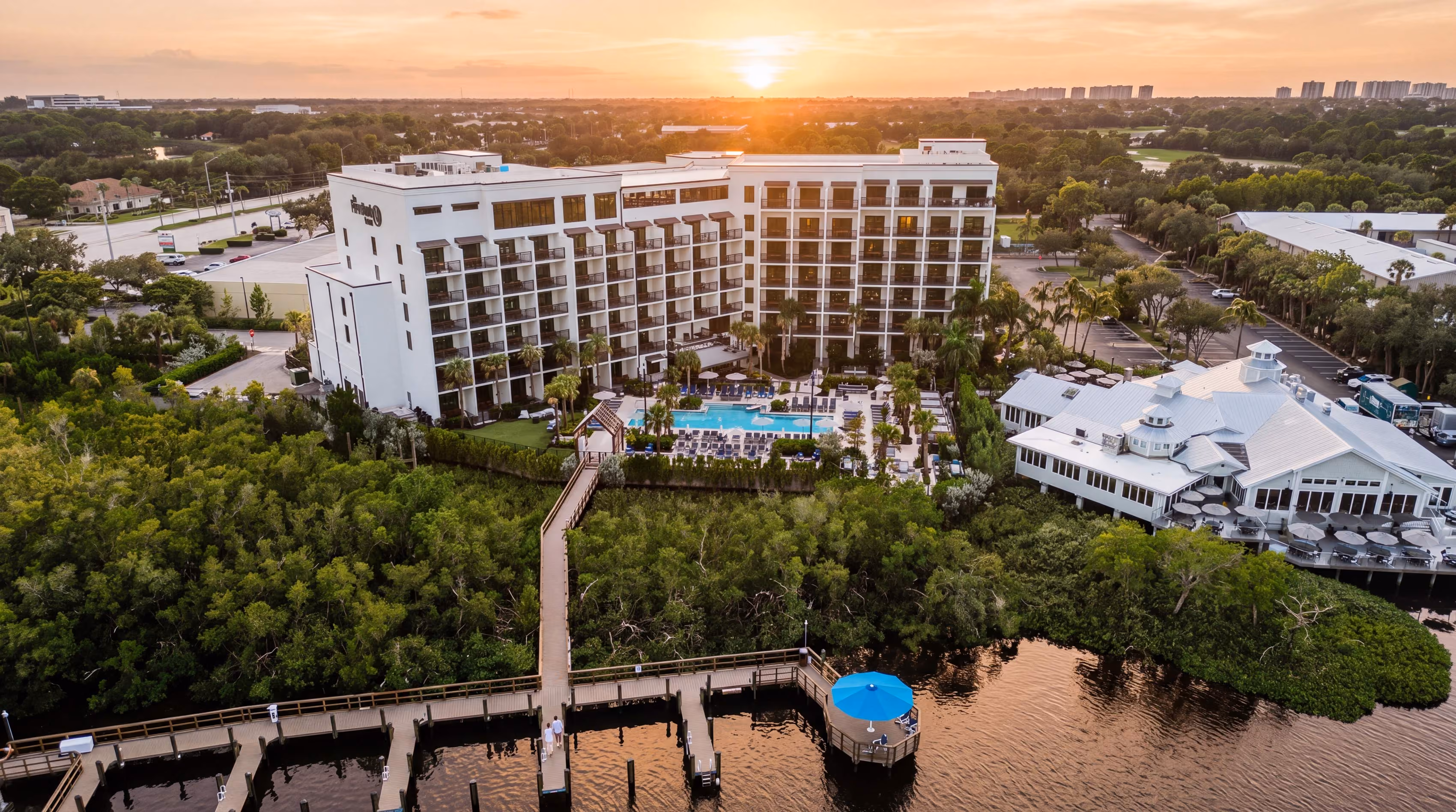 Aerial view of a large white hotel building with balconies surrounding a central outdoor swimming pool, next to a waterfront dock with a blue umbrella-covered seating area, surrounded by dense green trees, under a sunset sky.
