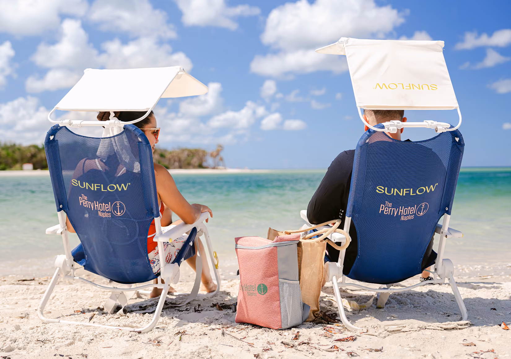 Couple sitting on blue Sunflow beach chairs with white canopies, facing a calm ocean on a sunny beach with clear blue sky and scattered clouds, with a red and beige beach bag placed between them on the sand.