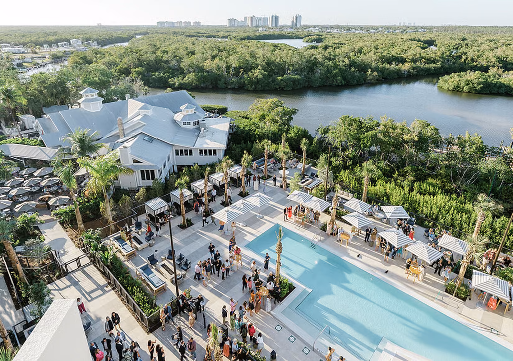 Aerial view of an outdoor event by a rectangular swimming pool surrounded by palm trees, striped umbrellas, cabanas, and groups of people socializing, with green trees and water in the background and a building to the left.