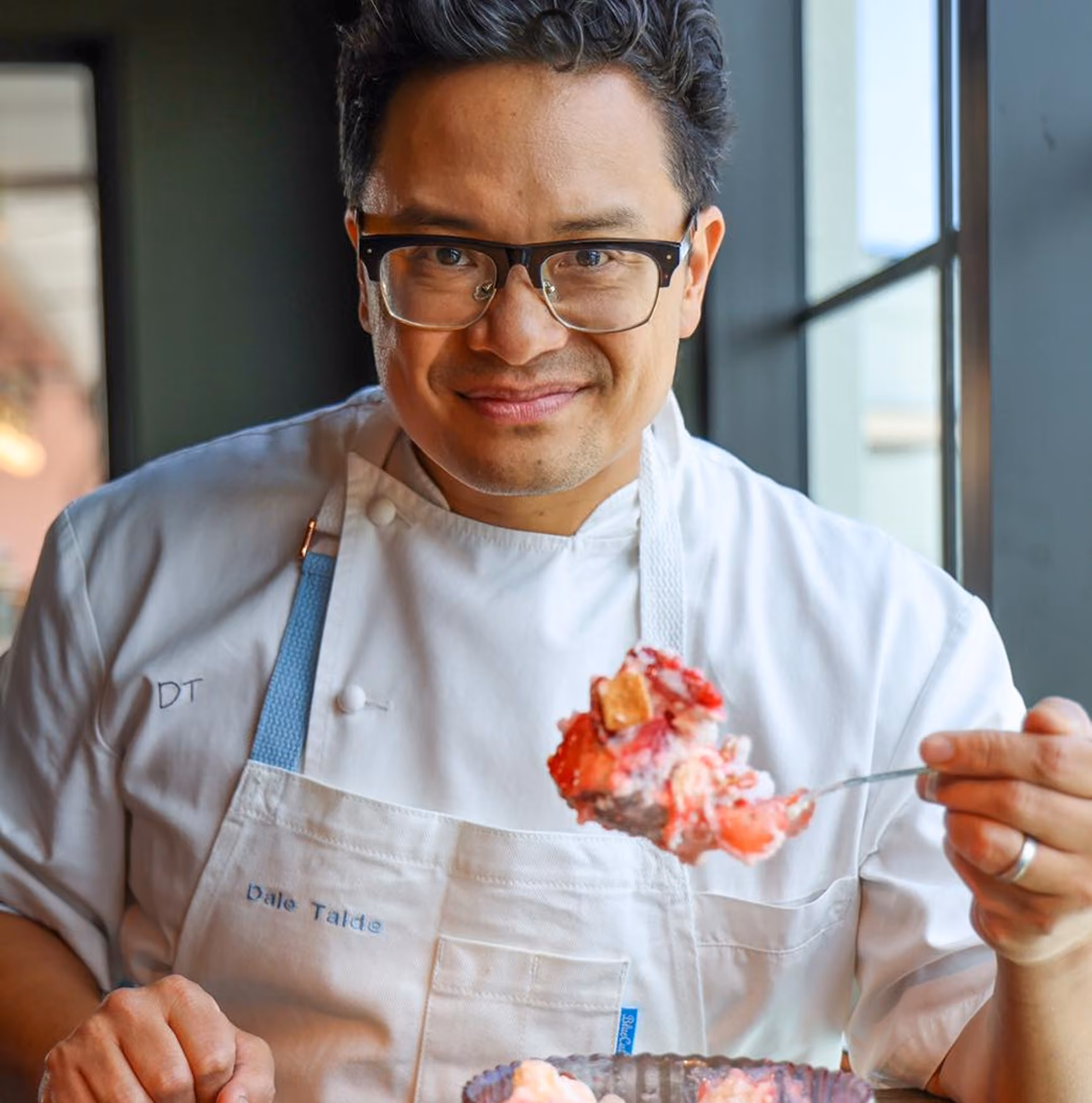 Chef Dale Talde wearing glasses and a white chef's coat with his name embroidered, holding a spoonful of dessert with strawberries and cream.
