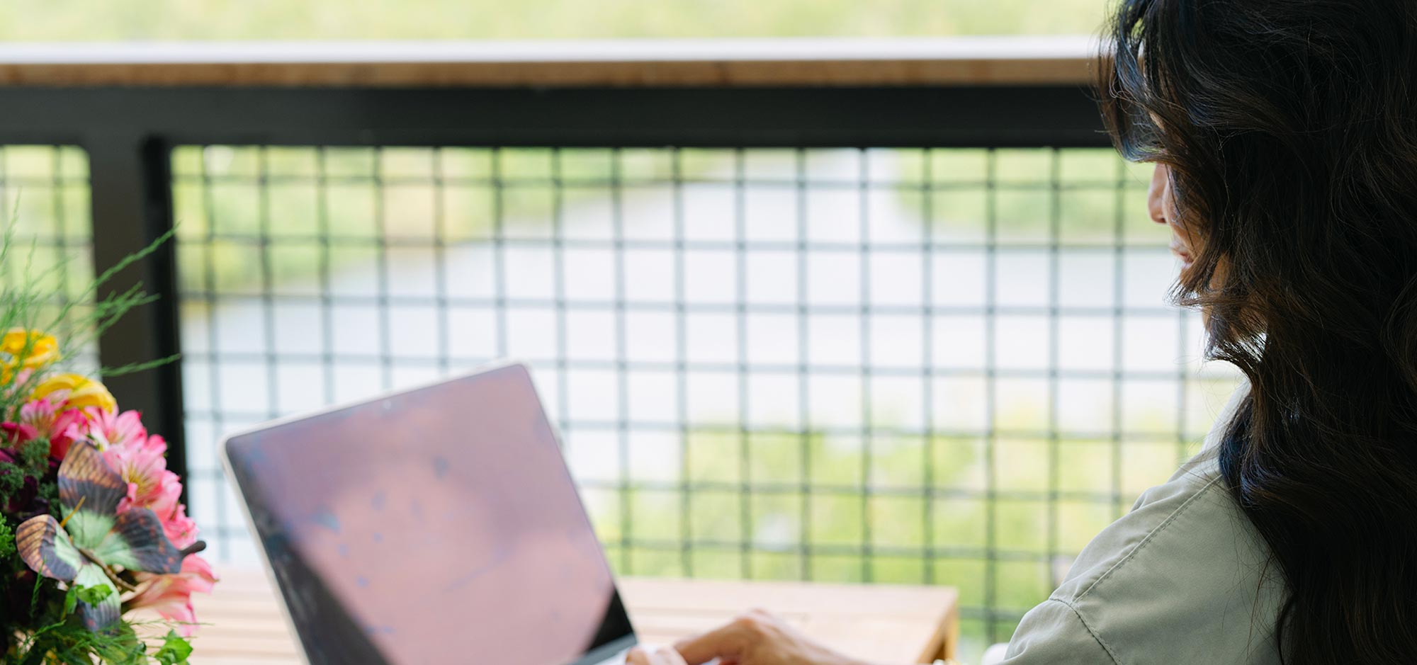 Woman with long dark hair sitting at a wooden table outdoors, looking at a laptop with a bouquet of flowers nearby, with a metal grid fence and greenery in the background.