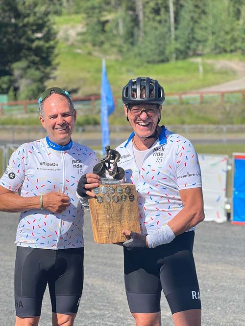 Two cyclists in matching white jerseys with colorful dots hold a trophy. They are smiling in a sunny outdoor setting, suggesting a sense of achievement.