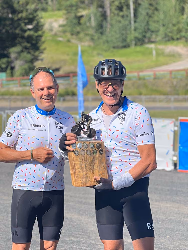 Two cyclists in matching white jerseys with colorful dots hold a trophy. They are smiling in a sunny outdoor setting, suggesting a sense of achievement.