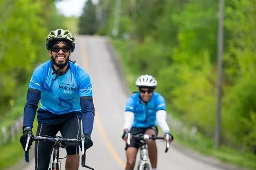 Two cyclists in blue jerseys ride on a rural road surrounded by greenery, smiling and enjoying the ride.