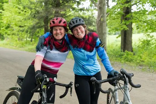 Two smiling cyclists with helmets and bright jerseys pose arm in arm beside their bikes on a leafy road, conveying friendship and joy.
