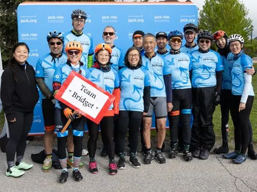A group of smiling cyclists in matching blue jerseys pose in front of a "jack.org" backdrop. One holds a sign reading "Team Bridget," conveying team spirit.