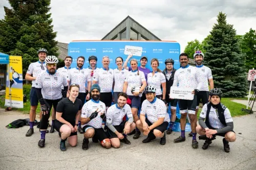 A group of cyclists in colorful jerseys pose smiling in front of a "jack.org" backdrop. Two hold signs. The mood is enthusiastic and celebratory.