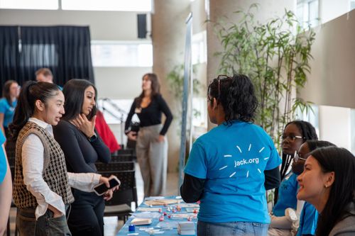 People engage at a booth, with one in a "jack.org" shirt facing the camera. The scene is lively, with conversations and informational materials visible.