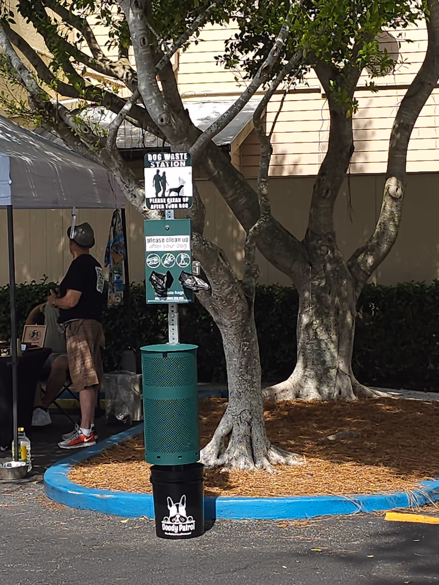 Dog waste station with a green trash bin, black bucket labeled Doody Patrol, and signs reminding owners to clean up after their dogs, set by a mulched tree area.