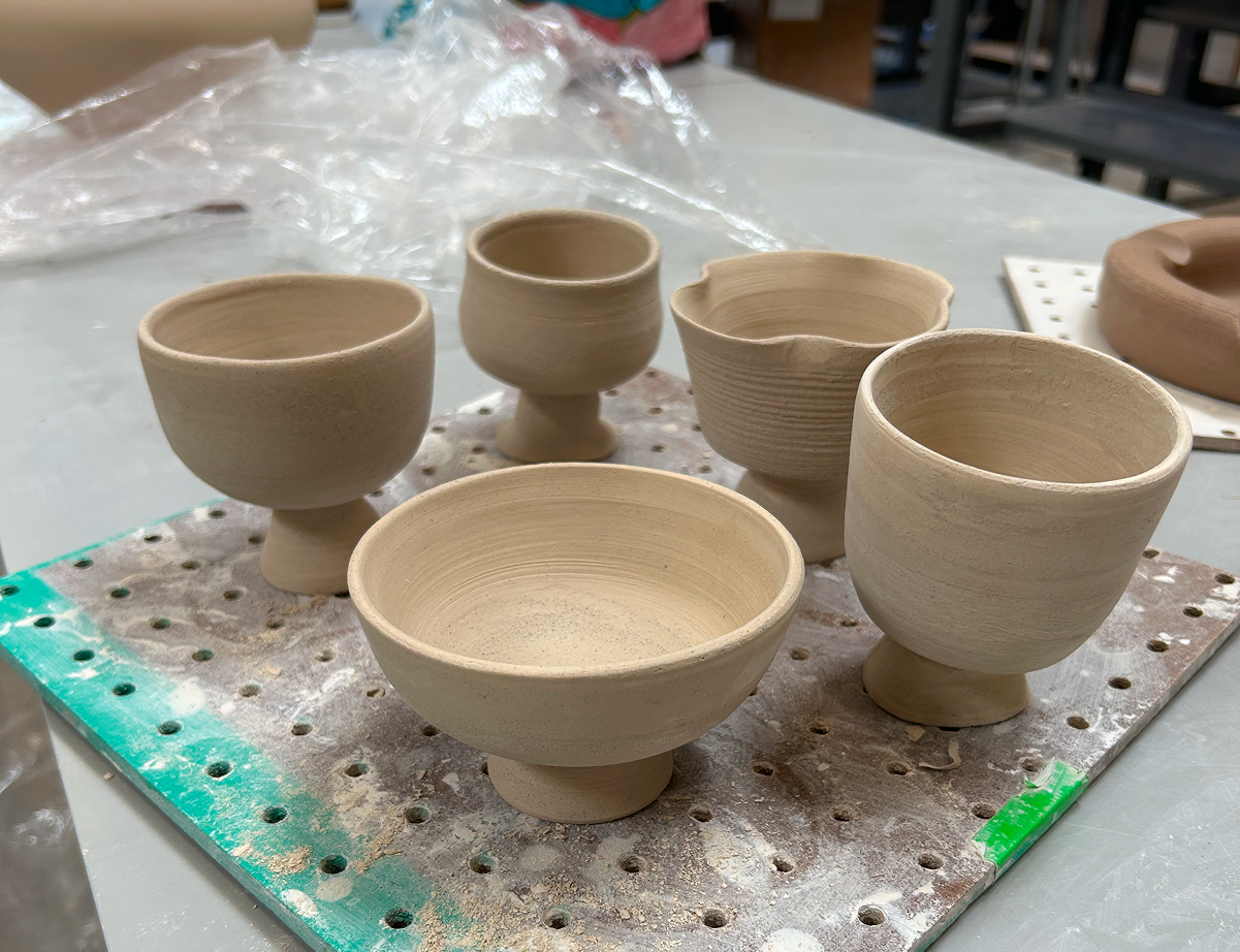 Five unglazed ceramic cups and bowls on a perforated board in a pottery studio.