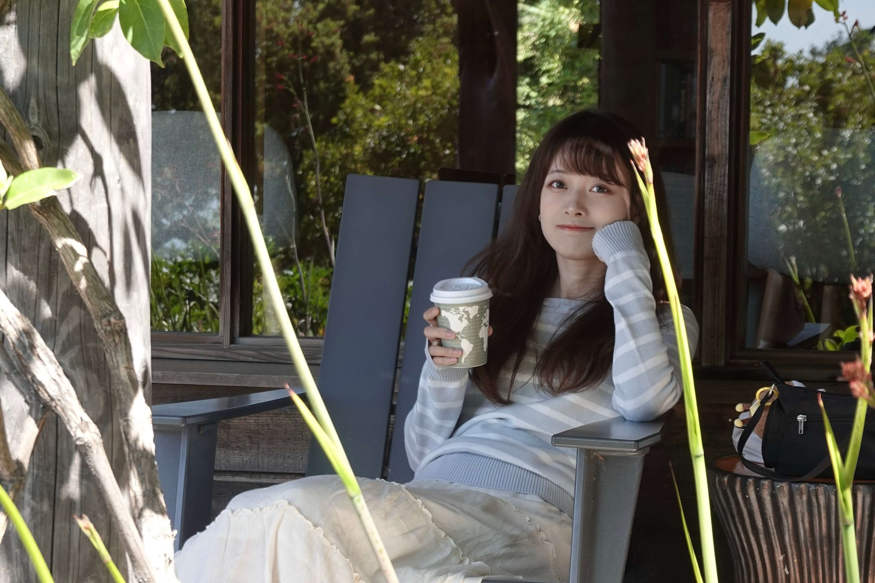 Young woman in a striped sweater sitting on a chair outdoors holding a paper cup and smiling.