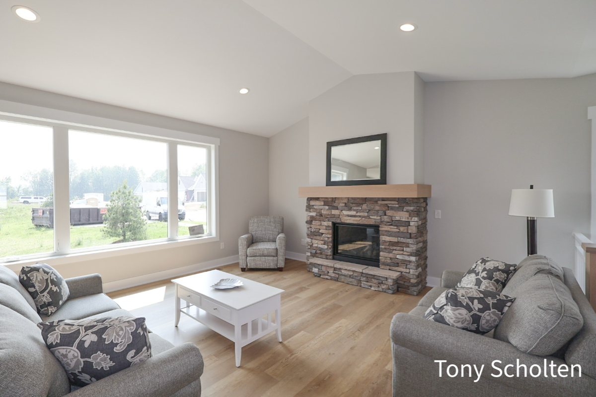 Bright living room with large window, two gray sofas with patterned pillows, a gray armchair, white coffee table, and stone fireplace with mirror above.