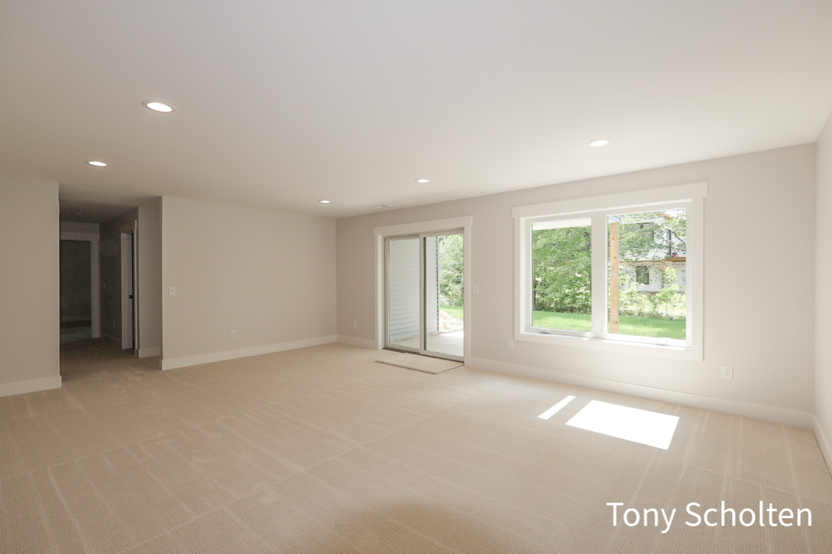 Bright unfurnished room with beige carpet, white walls, recessed ceiling lights, a sliding glass door, and a large window overlooking a green yard.