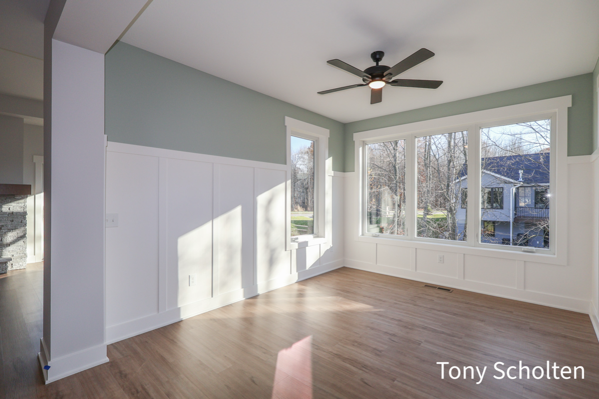 Unfurnished bright room with light wood flooring, gray-green upper walls, white paneling, large windows, and a black ceiling fan.