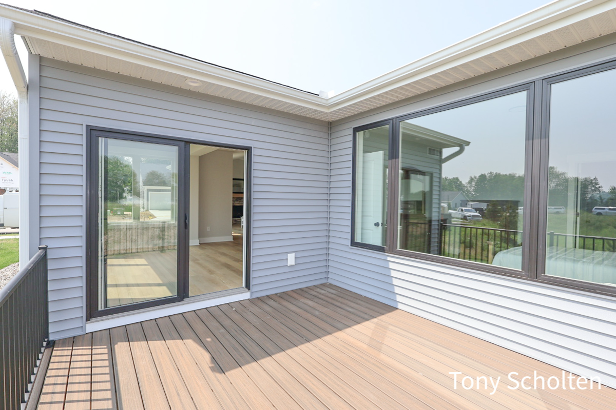 Outdoor corner deck with wood flooring, gray siding, sliding glass door, and large window reflecting a green yard