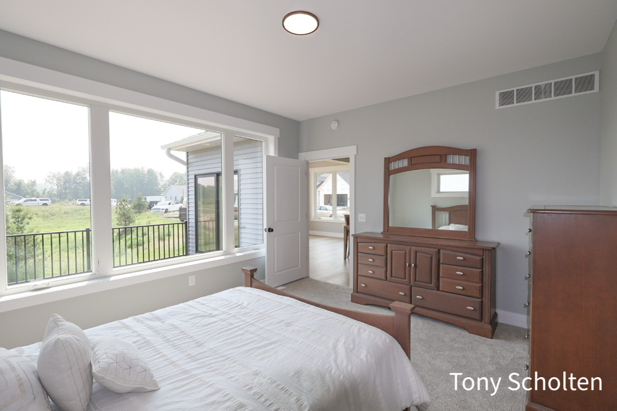 Bright bedroom with large windows, a wooden bed with white bedding, a wooden dresser with mirror, and a door open to another room.