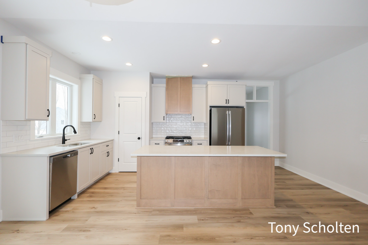 Modern kitchen with light wood flooring, white cabinets, a central island with light wood paneling, stainless steel appliances, and recessed ceiling lights.