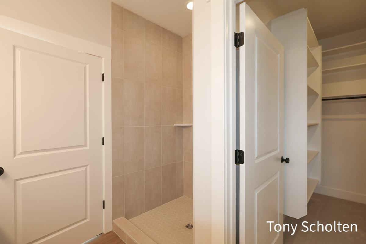 Bathroom with beige tiled walk-in shower and adjacent white door leading to a closet with shelves and hanging rods.