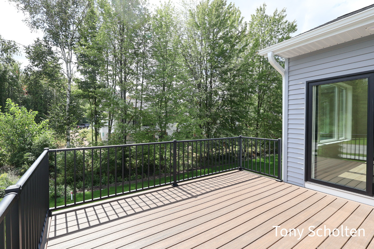 Unfurnished wooden deck with black metal railing overlooking lush green trees and a sliding glass door on a house with gray siding.