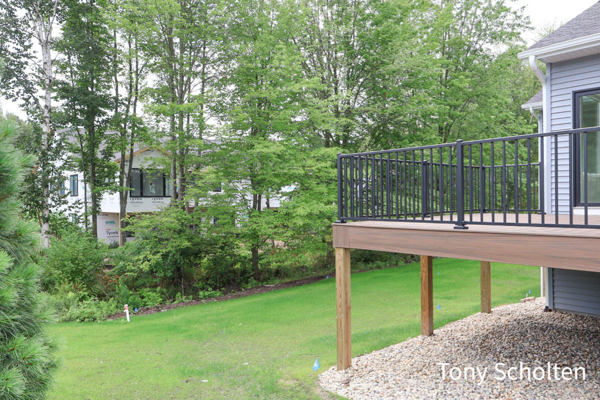 Backyard with green lawn, gravel bed beneath a raised deck with black railings, and a house exterior partially visible on the right.