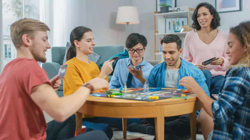 Gruppo eterogeneo di ragazzi e ragazze che giocano a un gioco da tavolo strategico con carte e dadi che leggono le carte e ridono. Amici che si divertono. Accogliente soggiorno durante il giorno.