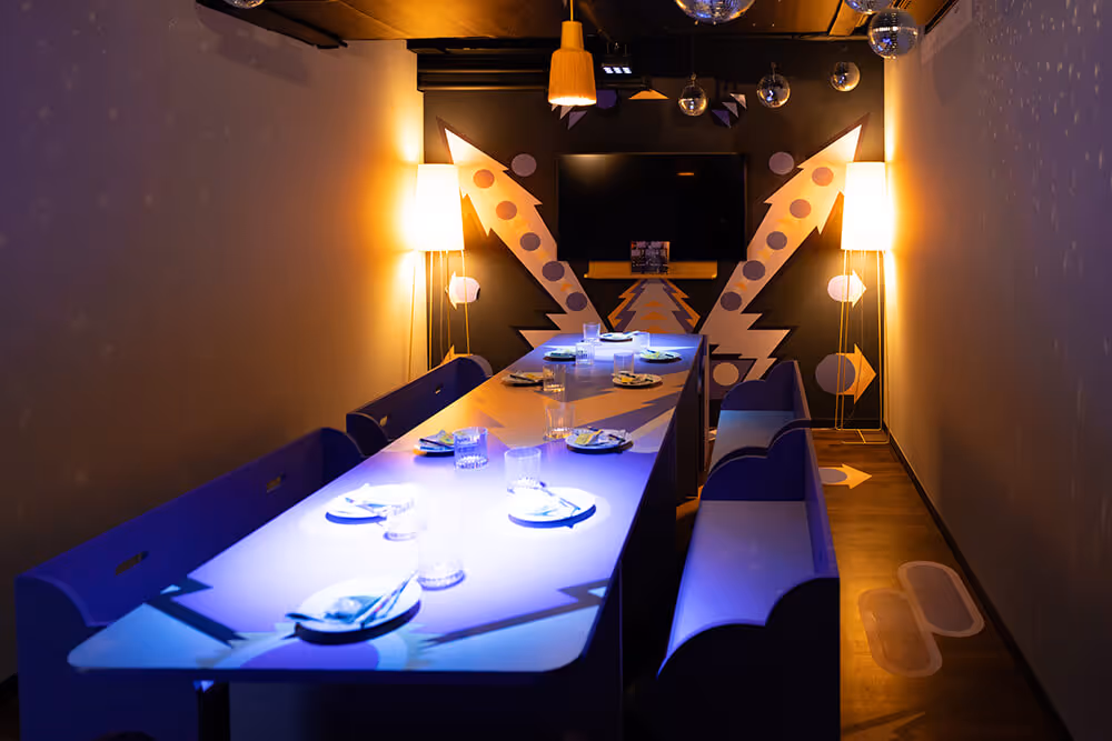 Moderner, stilvoll beleuchteter Konferenzraum mit einem langen Tisch, blauen Bänken, Lampen an beiden Seiten und einem Fernseher an der Wand.