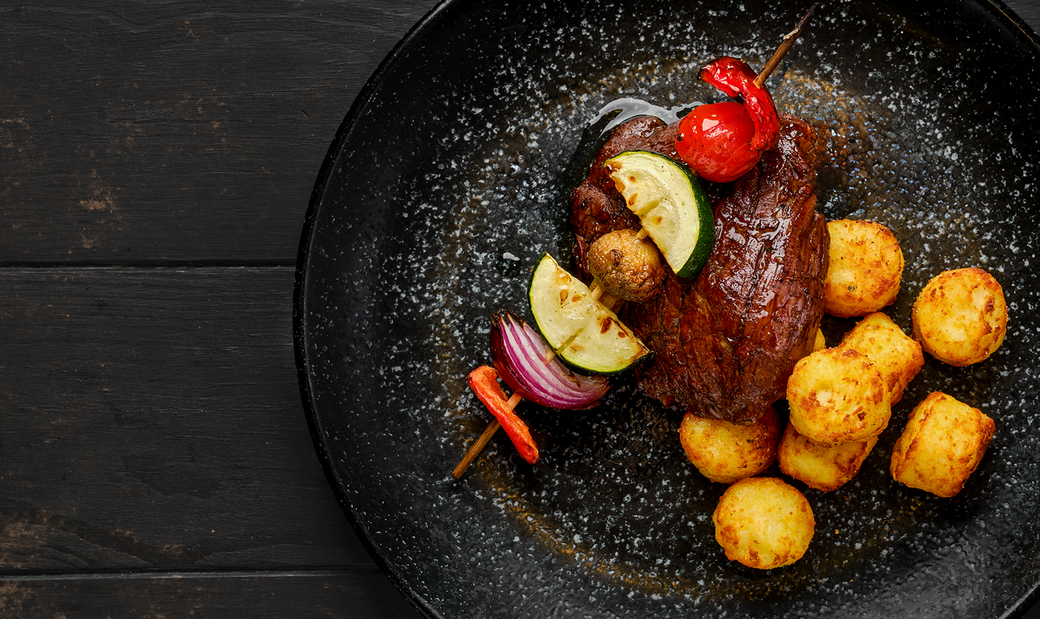 Grilled steak with vegetable skewer and crispy potato croquettes on a black plate.