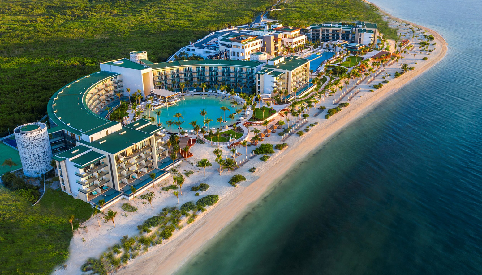 Aerial view of a large beachfront resort with a curved central building, swimming pool, palm trees, white sandy beach, and green forest in the background.