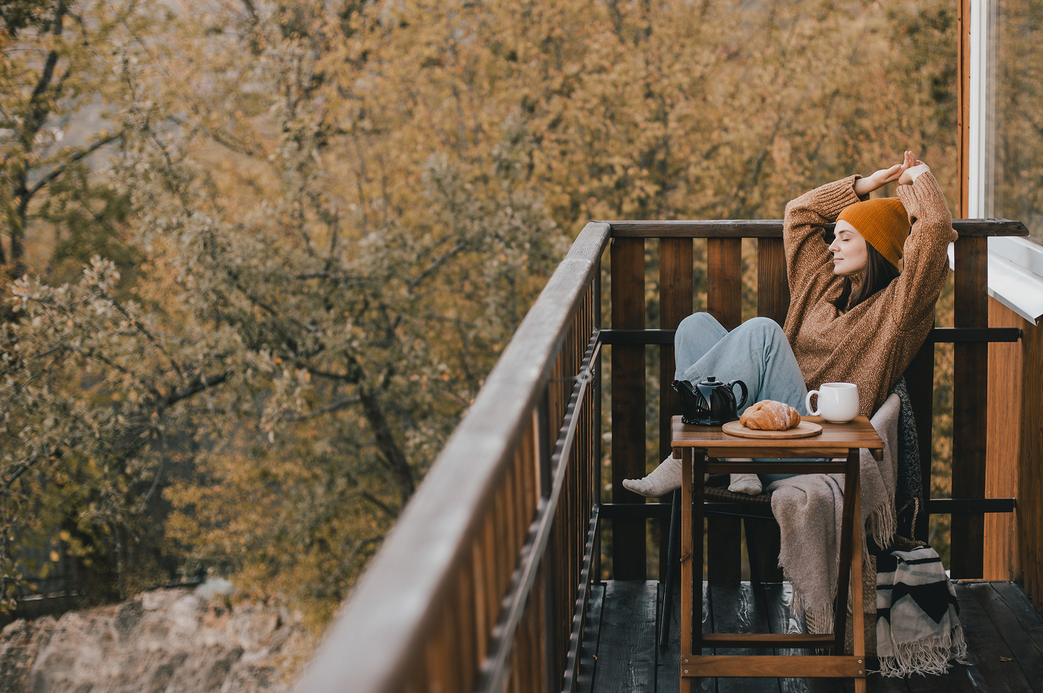Woman in a brown sweater and orange beanie sitting on a balcony with autumn trees in the background, enjoying tea and a croissant.