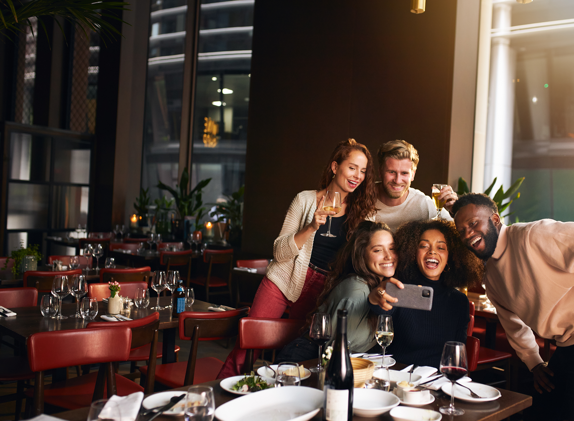 Group of five diverse friends smiling and taking a selfie together at a restaurant table with wine glasses and dishes.