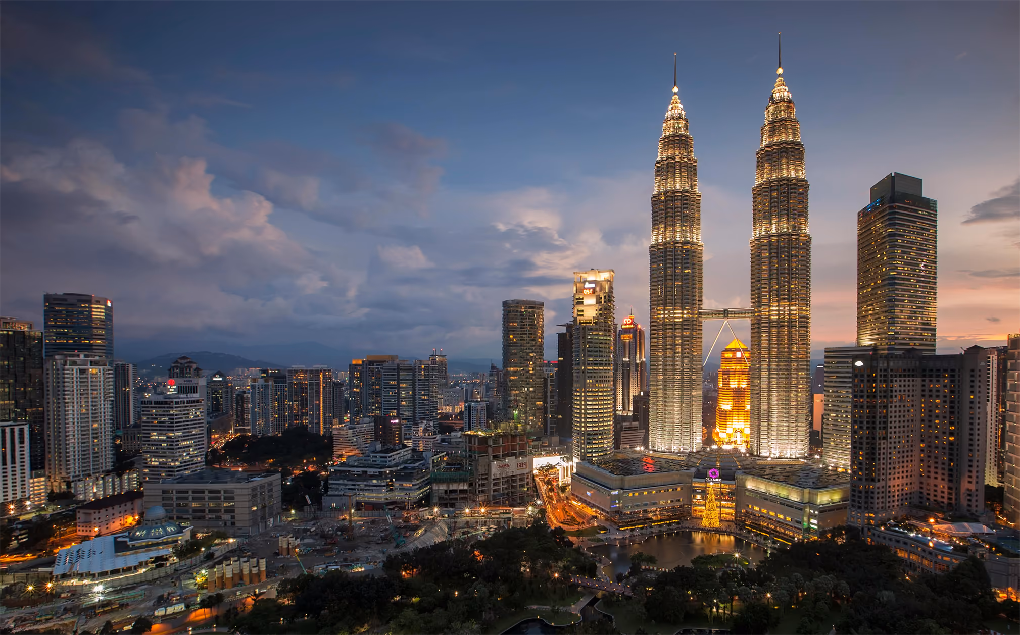 Night view of Kuala Lumpur city skyline featuring the illuminated Petronas Twin Towers and surrounding skyscrapers under a cloudy sky.
