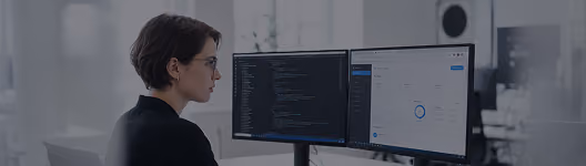 A woman with short hair and glasses working at a desk with dual computer monitors displaying code and charts in a modern office.