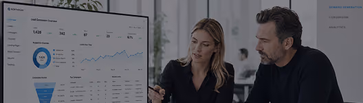 Two business professionals, a woman and a man, discussing data displayed on a large screen showing lead generation metrics, graphs, and charts in a modern office setting.